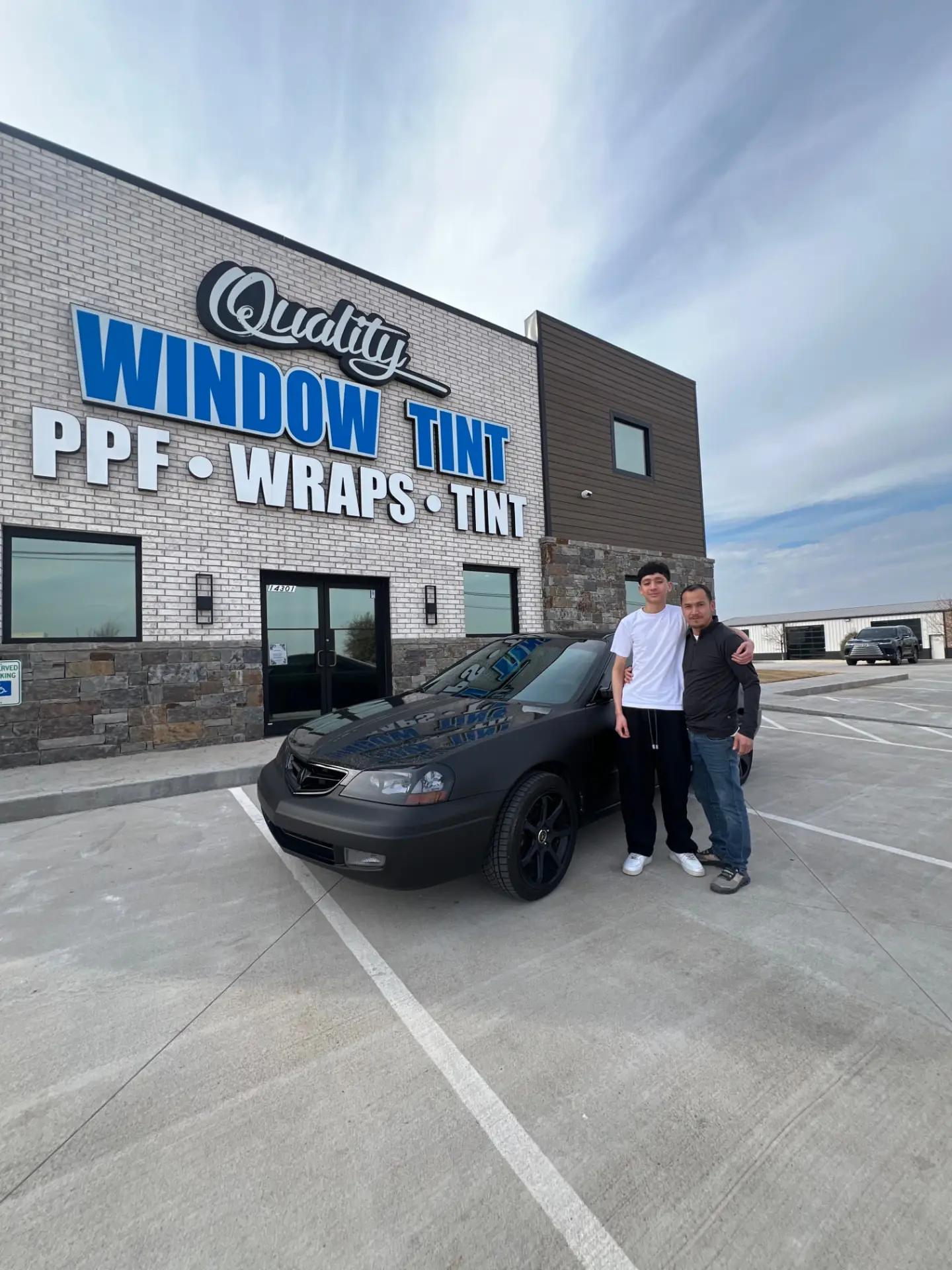 Two members of the Quality Window Tint team with a matte-black wrapped car in front of the Edmond shop